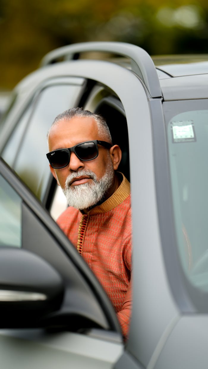 Confident bearded man in sunglasses posing in a car's driver's seat on a bright day.